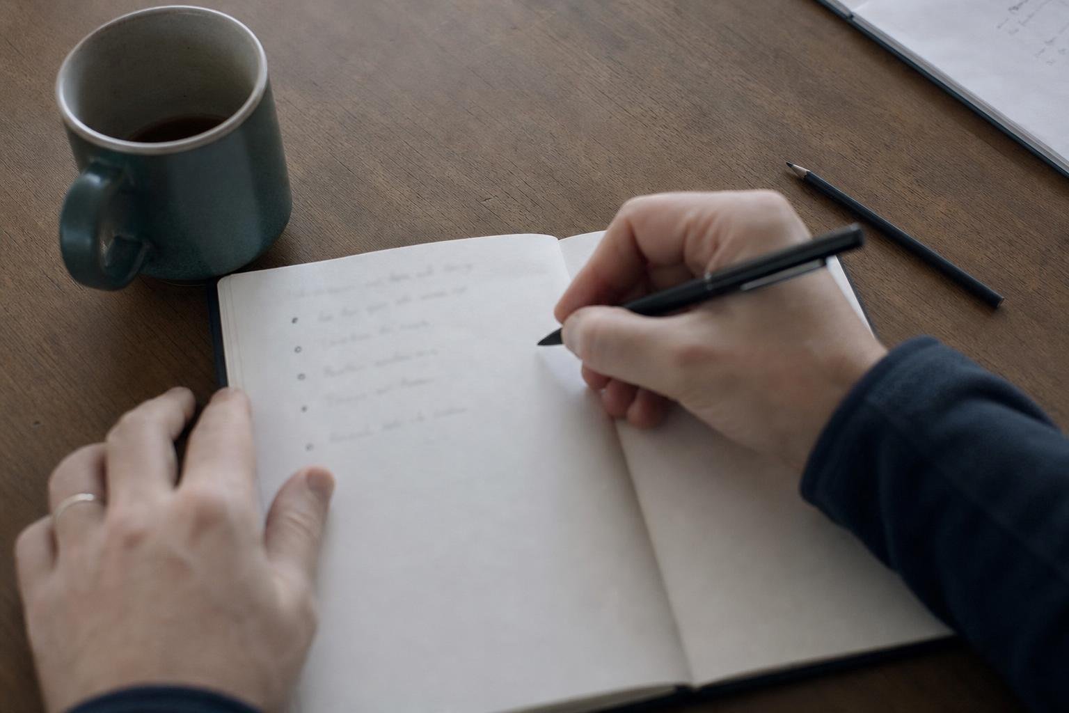 Hands writing an agency shortlist in a notebook beside a mug of tea