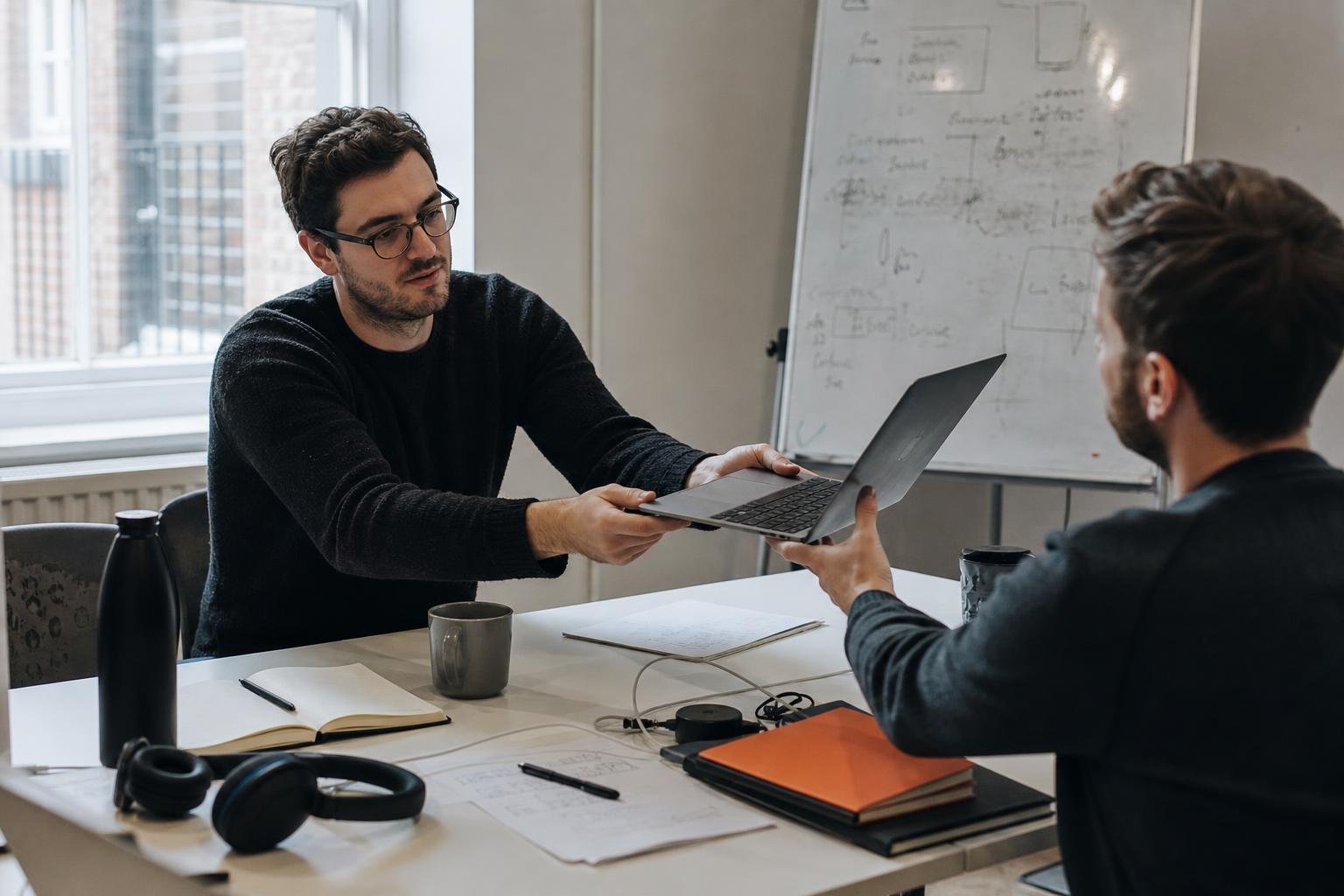 Two colleagues in a small office passing a laptop across a desk, the moment a SaaS PPC agency handover starts