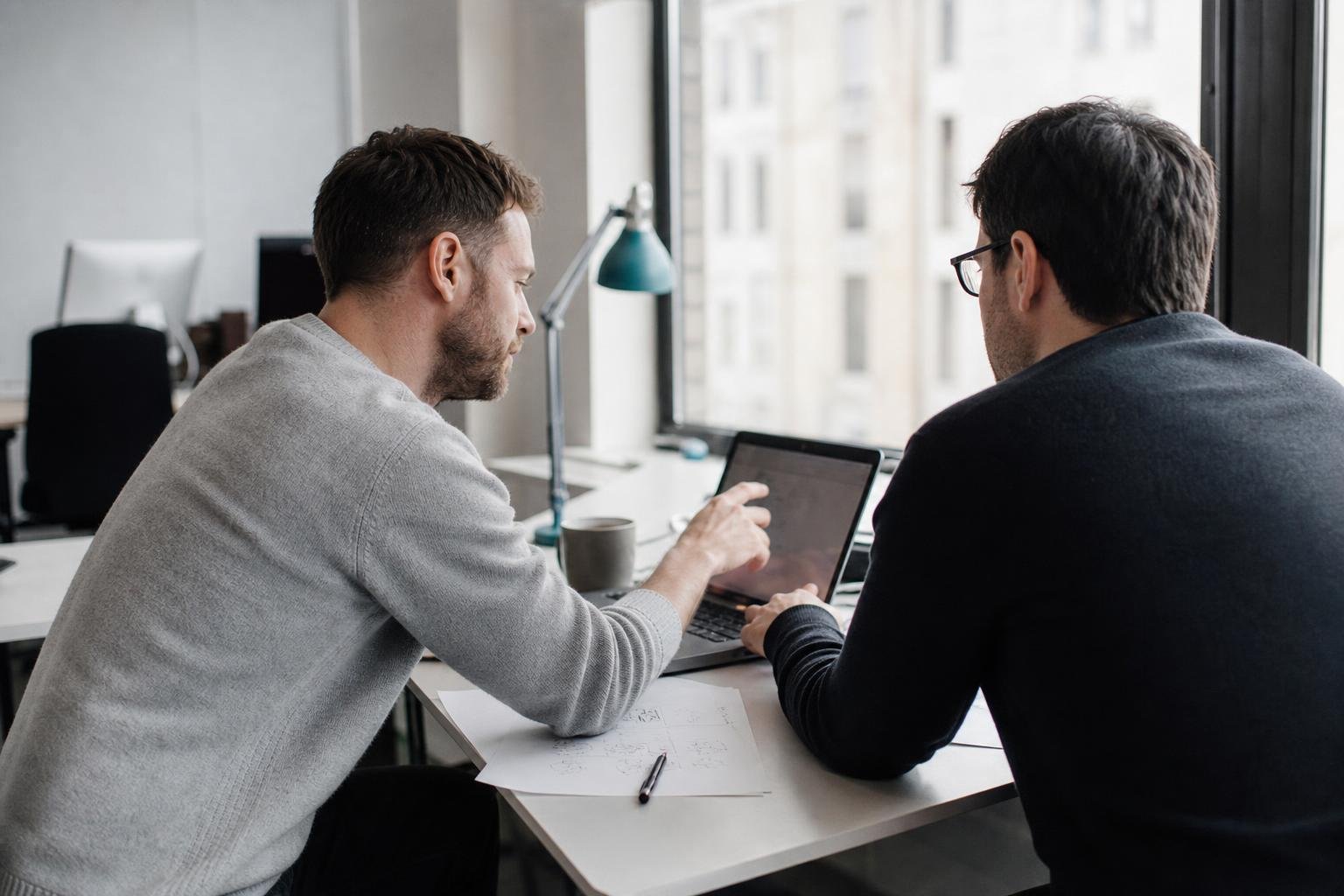 Two founders of a small SaaS company comparing Google Ads management tools on a laptop at a window desk