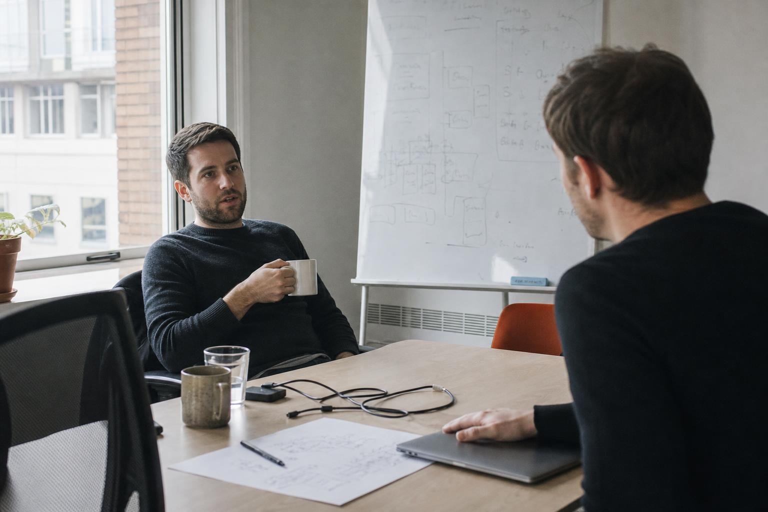 Two colleagues in a small UK office talking across a desk about hiring an ads agency or running the ads with software