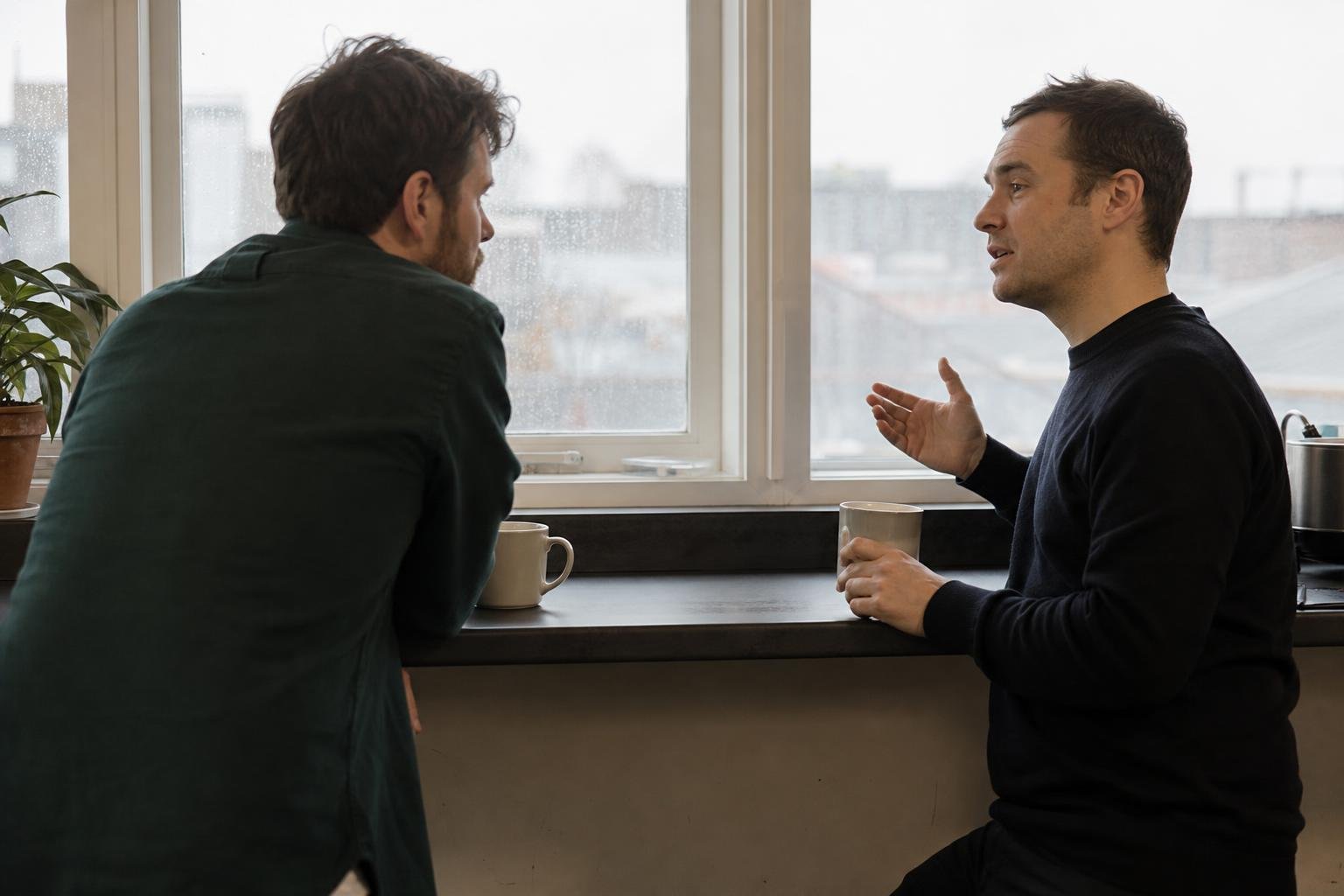 Two cofounders talk over mugs of tea at an office kitchen counter, rain on the window, deciding who should run their SaaS ads